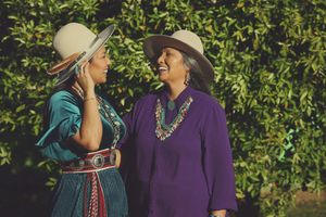 Two smiling women in wide-brimmed hats and colorful embroidered western outfits laugh together in front of a leafy green hedge.