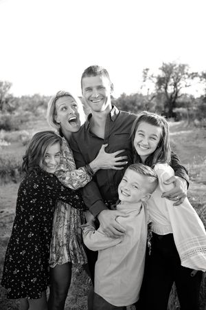 A black and white portrait of a laughing family of five hugging closely outdoors, with a dad, mom, two girls and a young boy.