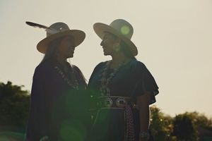 Two women in wide-brimmed hats, one with a feather, stand face to face in silhouette against a soft golden sky at sunset.