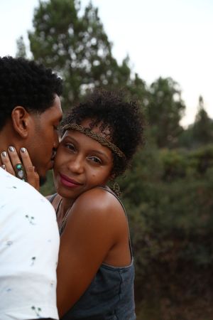 A woman in a braided headband smiles softly at the camera as a man kisses her cheek, with green trees in the background.