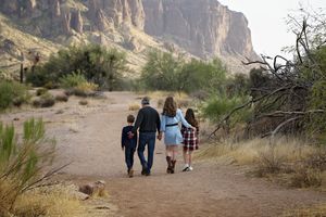 A family of four walks together along a desert trail toward rocky mountain cliffs and green desert shrubs.