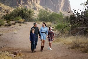 A family of four holds hands and walks along a sandy desert path with rugged mountains in the background.