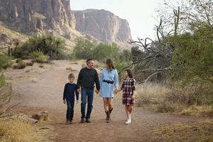 Parents and two children walk hand in hand down a desert trail beneath towering rocky cliffs.