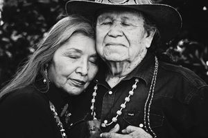Black and white portrait of an elderly couple embracing, the woman resting her head on a man in a wide-brimmed hat and beaded necklaces.
