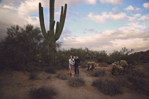 A family stands together near a tall saguaro cactus in the desert under a colorful sunset sky.