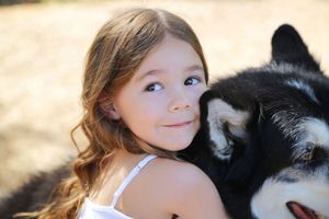 Smiling young girl with long wavy hair hugs a black and white husky dog outdoors in warm sunlight.