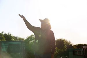 Person in a hat raises one arm toward the sky as bright sunlight flares behind them near green trees.