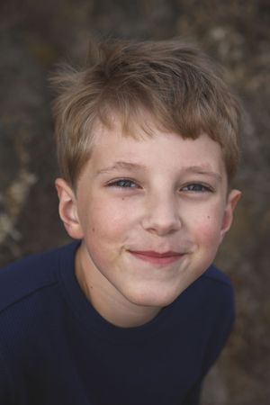 Smiling young boy with short blond hair and a navy shirt posing for a close-up portrait outdoors.