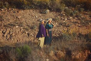 Two women in wide-brimmed hats and traditional-style clothing walk along a rocky hillside dotted with desert shrubs in warm light.