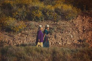 Two women in hats and colorful traditional dress walk along a rugged hillside surrounded by golden autumn shrubs.