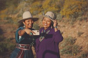 Two laughing women in felt hats and silver jewelry take a selfie with a smartphone on a golden desert hillside.