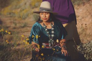 Woman in a felt hat, teal velvet blouse, and large silver squash blossom necklace sits among yellow wildflowers on a hillside.