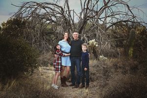 Family of four in coordinated outfits posing together in a desert landscape beside a gnarled bare tree and cactus.