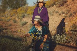 Two women in hats and silver turquoise jewelry pose on a sunlit hillside, one standing behind the other among yellow wildflowers.