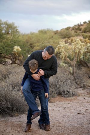 Father in a black sweater playfully hugging his young son from behind on a desert trail with cactus and shrubs.