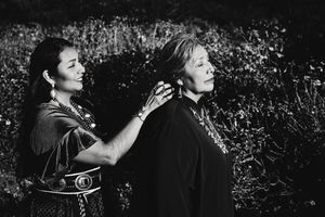 Black and white portrait of two women in traditional attire, one standing behind the other touching her shoulders, among wildflowers.