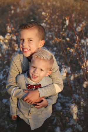 Two smiling brothers hugging in a cotton field at sunset, the younger wearing a red bow tie and gray button-up shirt.
