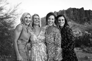 Black and white photo of four laughing women embracing in front of a rugged desert mountain landscape.