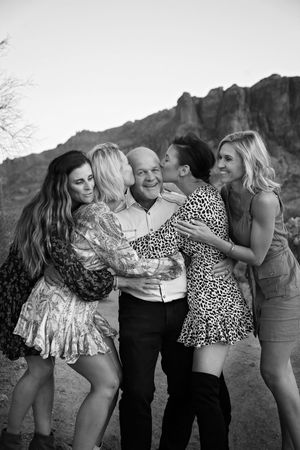 Black and white photo of four women hugging and kissing a smiling older man outdoors with desert mountains behind them.