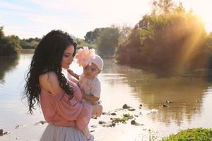 Mother with long dark curly hair holding her baby in a pink flower headband beside a sunlit lake with golden light.
