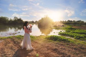 Woman in a flowing white dress holding a baby beside a calm lake as golden sunlight breaks through the clouds.