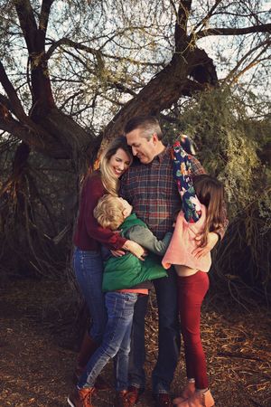 A smiling family of four shares a warm group hug beneath a large tree during a golden hour outdoor portrait session.