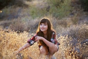 Smiling young girl with long brown hair in a plaid shirt crouching in golden grass during warm evening light.