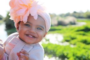Smiling baby wearing a large peach flower headband and cozy knit outfit in front of green grass and water.