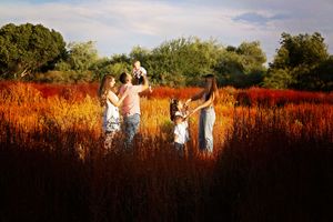 Family of five standing in a vibrant orange and red field at sunset, with parents lifting a baby and children holding hands.