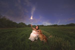 Woman in a white dress kneeling with her golden retriever in a grassy field at dusk, holding a glowing sparkler under a purple sky.