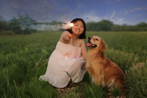 Smiling woman in a pale dress holding a sparkler beside her golden retriever in a green meadow at twilight.