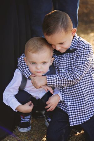Two young brothers in checkered shirts and bow ties hugging closely in an outdoor family portrait.