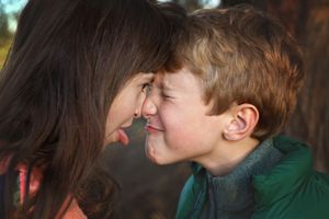 A girl sticks out her tongue as she touches foreheads with a grimacing boy in a green jacket outdoors.