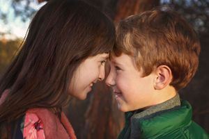 A smiling girl and boy touch foreheads in warm golden light outdoors, sharing a playful sibling moment.