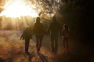A family of four holds hands and walks down a dirt path toward the glowing sunset, silhouetted by streaming sunlight.
