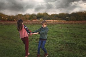 A girl and boy hold hands and spin together in a green grassy field beneath a dramatic cloudy autumn sky.