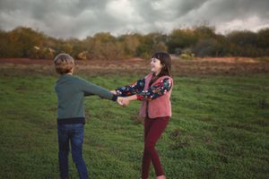 A brother and sister hold hands and laugh while spinning in a grassy meadow under moody gray clouds.