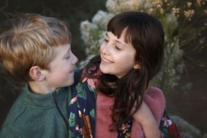 A smiling girl with long dark hair hugs her red-haired brother outdoors in front of soft flowering shrubs.