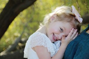 Smiling young girl with a pink bow in her blonde hair leans her head on her hand while sitting outdoors near a tree.