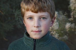 Close-up portrait of a red-haired boy with blue-green eyes wearing a green fleece jacket, softly lit outdoors.