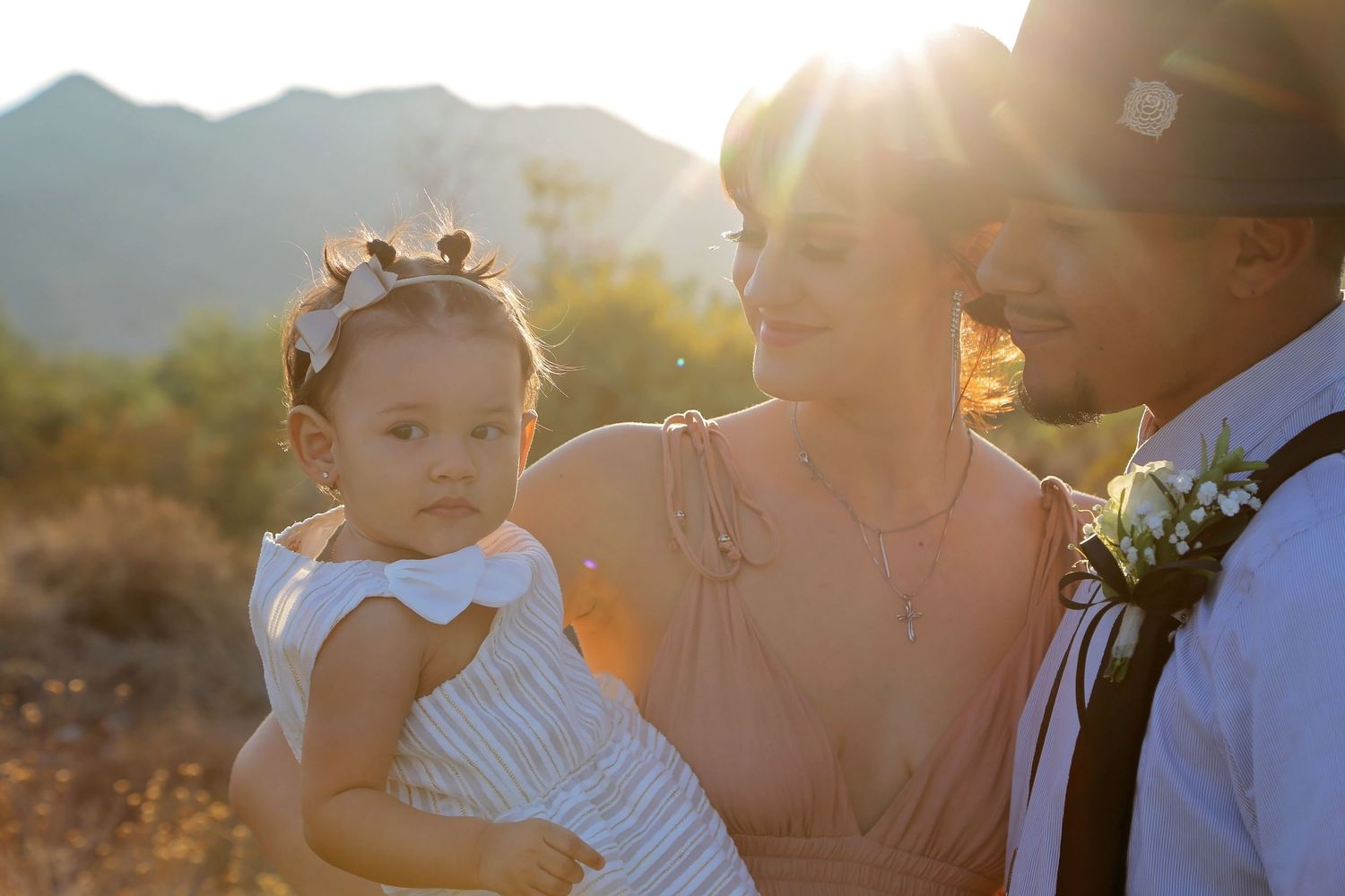 A toddler in a white dress and floral headband is held by her mother in glowing sunlight, with a man in a boutonniere nearby.