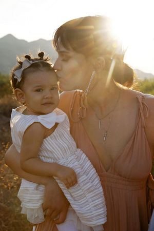 A smiling mother in a peach dress kisses her baby girl in a white striped dress outdoors in warm sunset light.