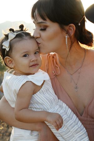 A mother in a blush dress and cross necklace lovingly kisses her baby girl wearing a bow headband and white dress.