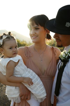 A happy family of three poses outdoors at golden hour, with a mother holding her baby girl beside a man in a black hat.