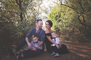 Mother, father and two young sons sit close together on a shaded dirt path surrounded by trees in a family portrait.