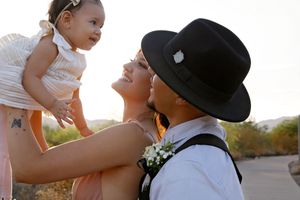 A smiling mother and father in a black hat gaze lovingly at their baby girl, held up in the warm desert sunlight.