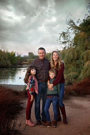 A family of four poses together by a pond at dusk, with parents hugging their two children under a cloudy sky.
