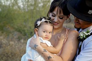 A baby girl in a white bow headband rests in her mother's tattooed arms as her father in a hat smiles beside them.