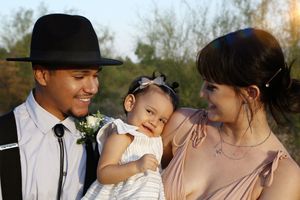 A smiling father in a black hat and boutonniere and a mother in a peach dress admire their giggling baby girl outdoors.