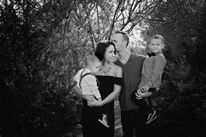 Black and white family portrait of a mother and father holding two young sons while standing among leafy trees.